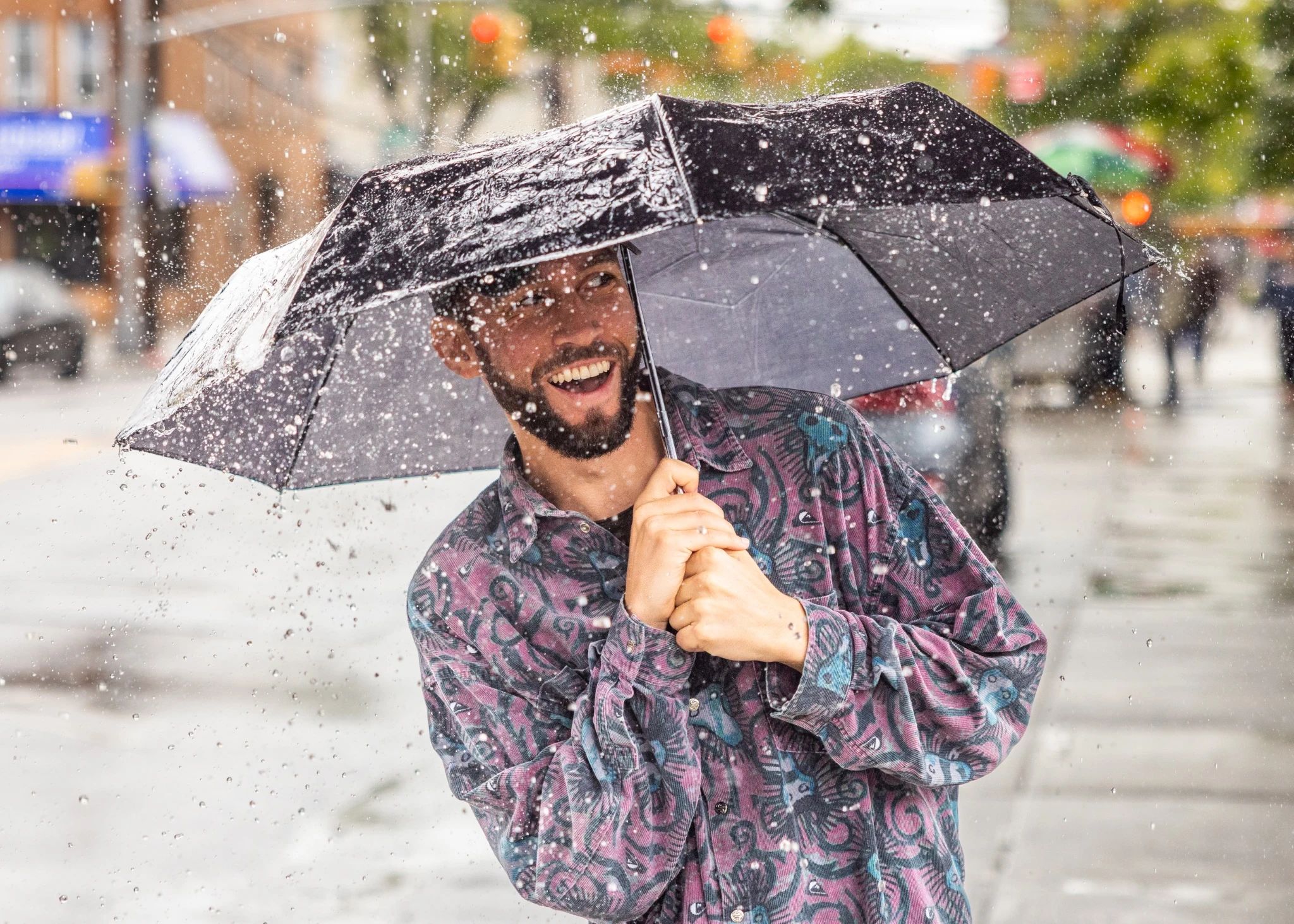 A smiling man holding an umbrella as a deluge of rainwater falls upon him.