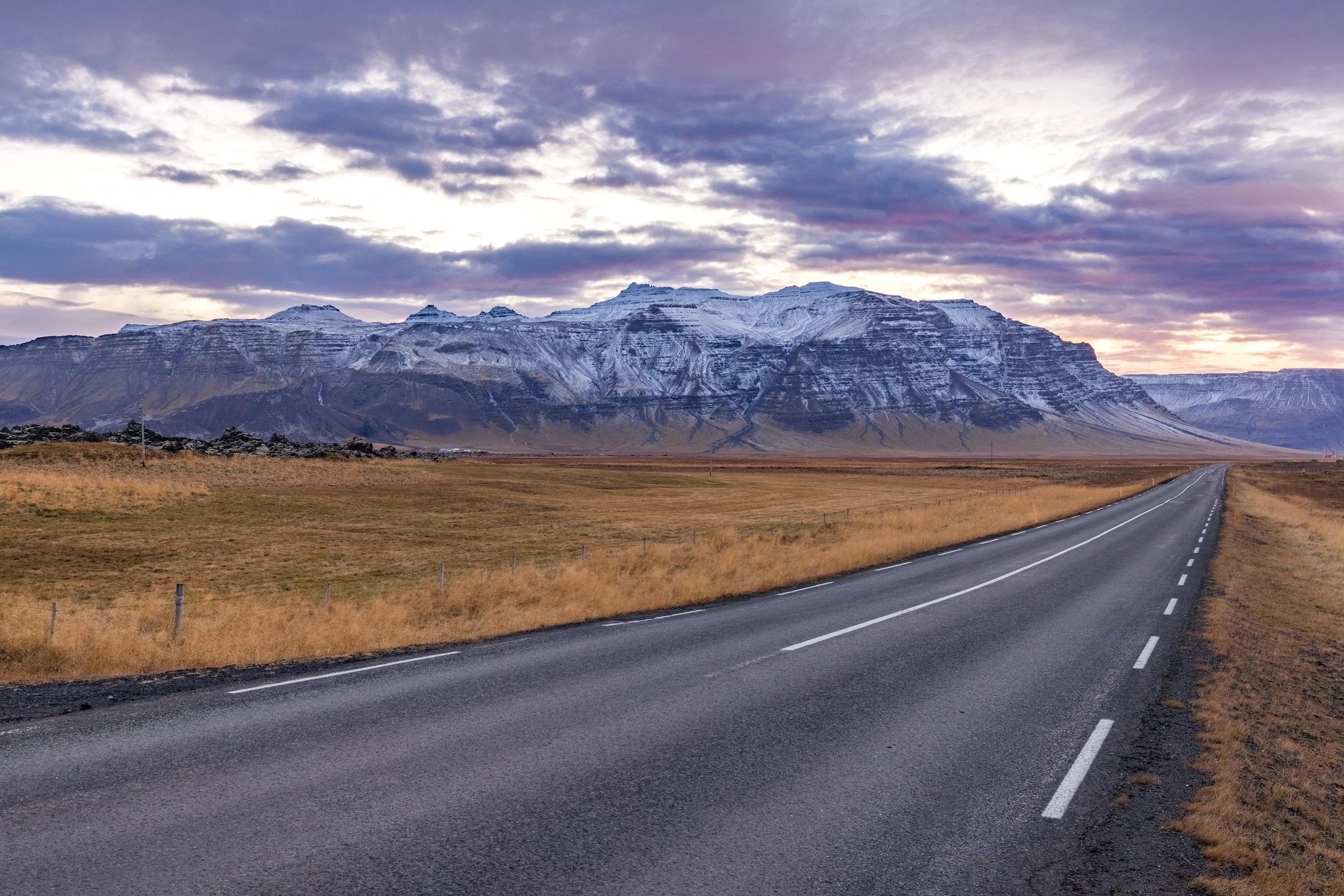 An Icelandic road stretching into the distance. In the background is a huge, snow-capped mountain.