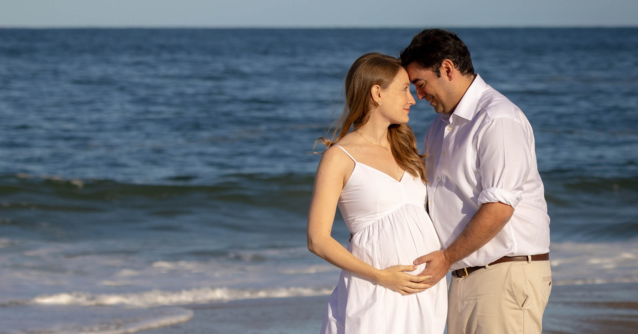 Two people standing on the beach, foreheads touching, looking into each others' eyes. The woman on the left is pregnant!