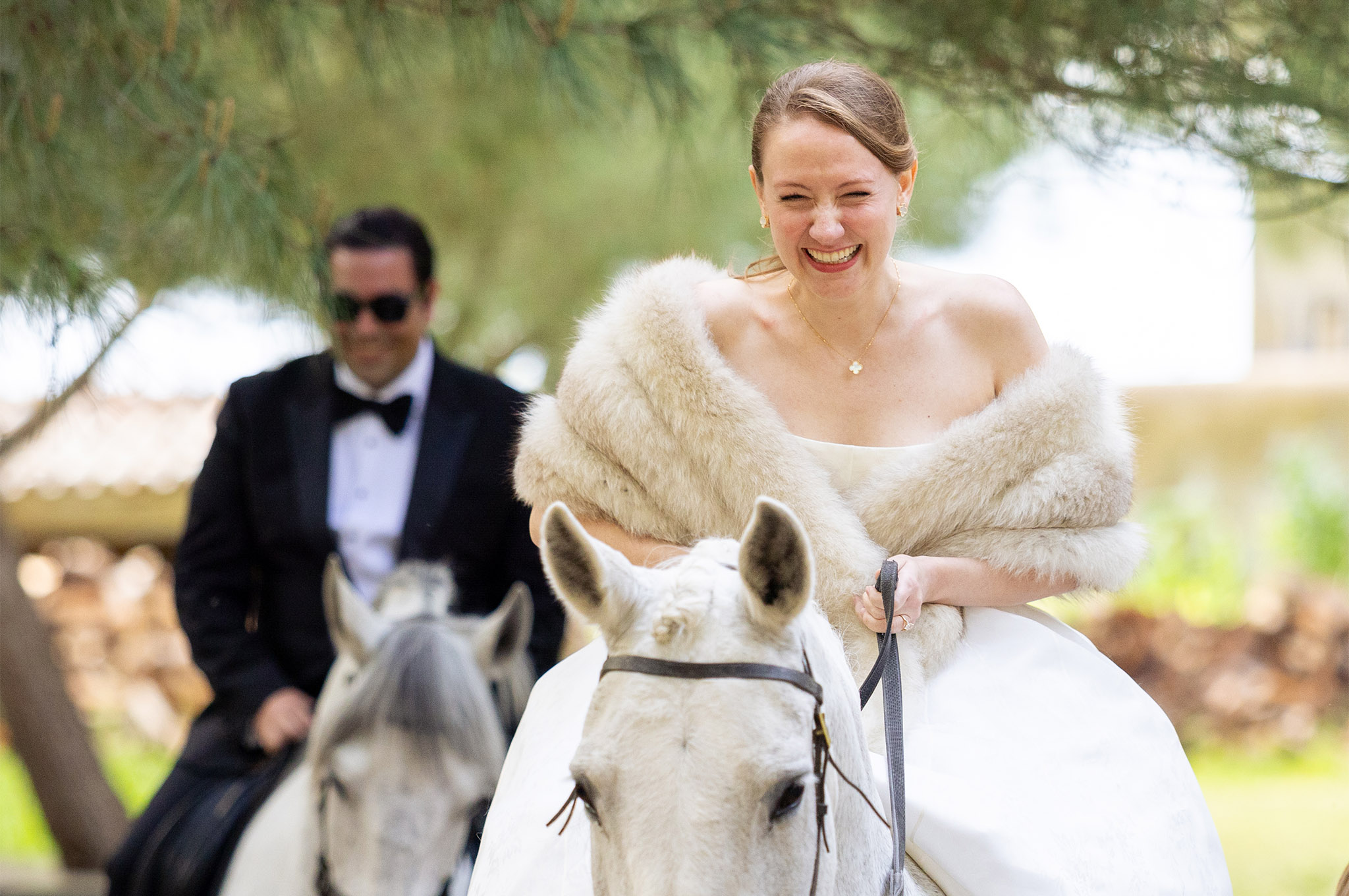 A married couple sitting on two separate horses. The bride is beaming in the foreground on the right side of the photo.