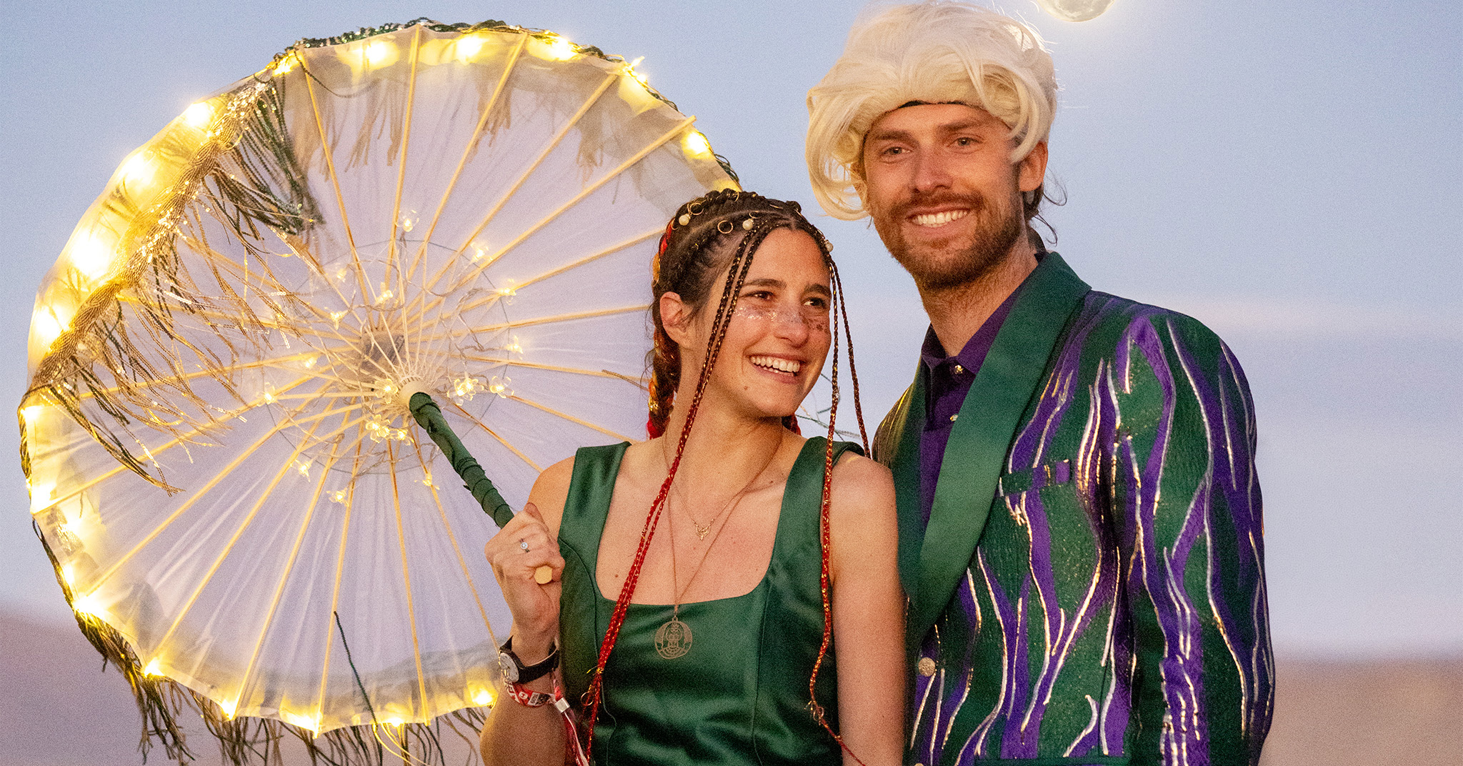 A woman and man dresssed in green and purple, smiling. The woman is holding a lit parasol.
