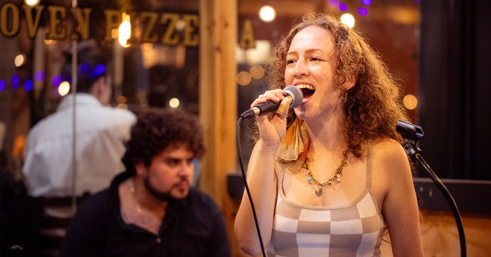 Mikayla Cohen singing "If I Ain't Got You" into a microphone in front of a pizzeria window in New York City.