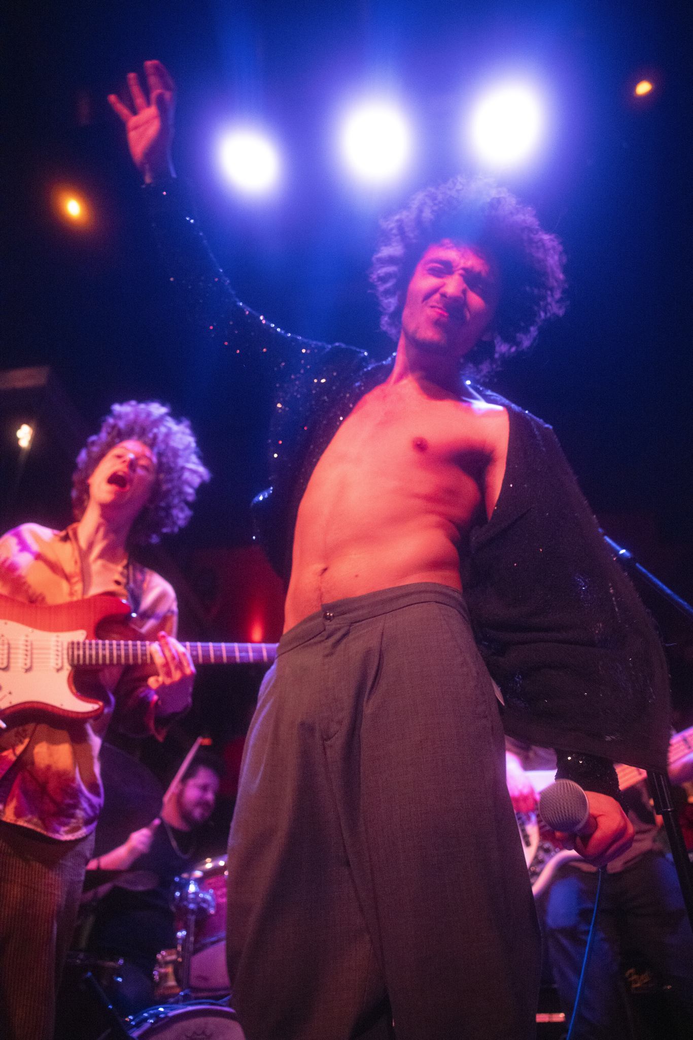 A musician reaching up towards lights while a guitarist shreds in the background