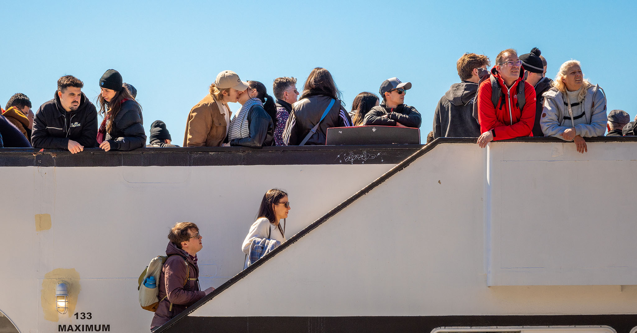 Several people are standing at the edge of the top deck of a Ellis Island ferry. Two people are walking up some stairs to get to the top deck. Two people are kissing each other on the top deck.