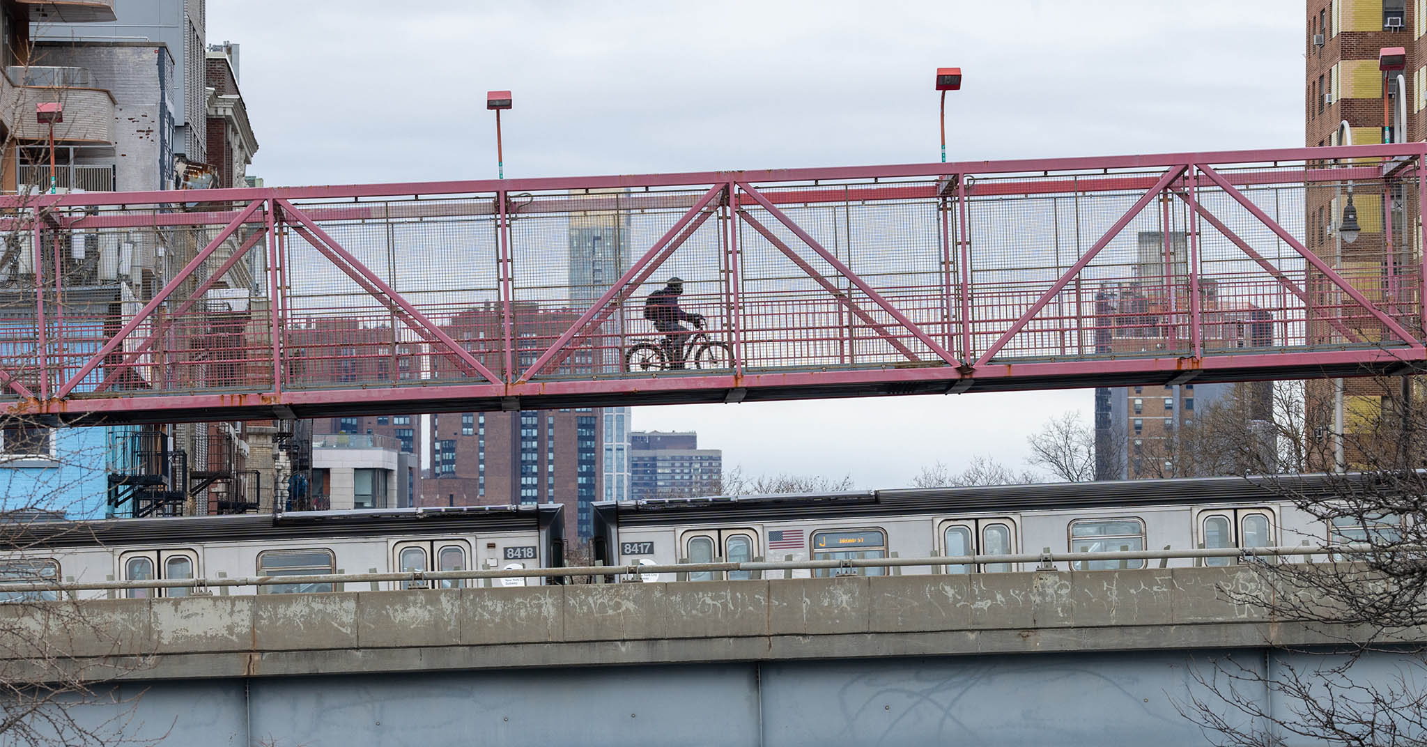 A cyclist is moving from left to right over the Williamsburg Bridge. A J subway train is moving below them.